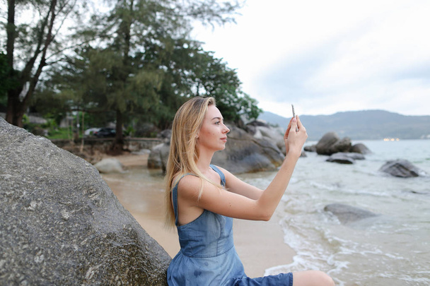 Молодая блондинка фотографируется на смартфоне и сидит на песке возле камней и моря
. - Фото, изображение