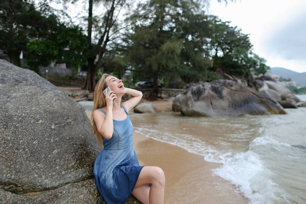 Молода красива дівчина сидить на піску біля моря і каменів, розмовляє по смартфону і показує великі пальці вгору
. - Фото, зображення