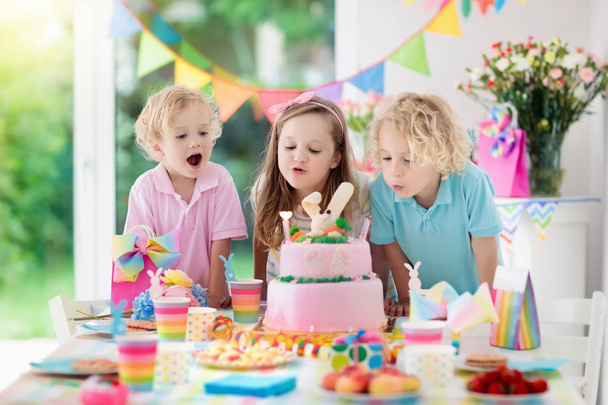 Kids verjaardagsfeest. Kinderen blazen kaarsjes op de taart roze konijntje. Pastel rainbow decoratie en tabel instellen voor kinderen gebeurtenis, banner en vlag. Meisje en jongen met verjaardagsgeschenk. Familiefeest. - Foto, afbeelding