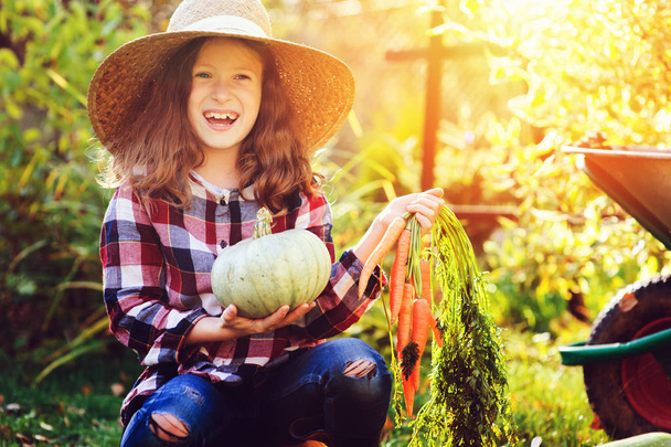 happy funny child girl in farmer hat and shirt playing and picking autumn vegetable harvest in sunny garden. Growing organic carrots and pumpkins. - Foto, immagini