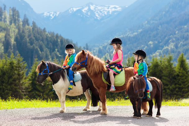 Kinder beim Ponyreiten in den Alpen. Familienfrühlingsurlaub auf einer Pferderanch in Österreich, Tirol. Kinder reiten auf Pferden. Kind kümmert sich um Tiere. Kind und Haustier. Mädchen und Junge im Sattel auf Pony. - Foto, Bild