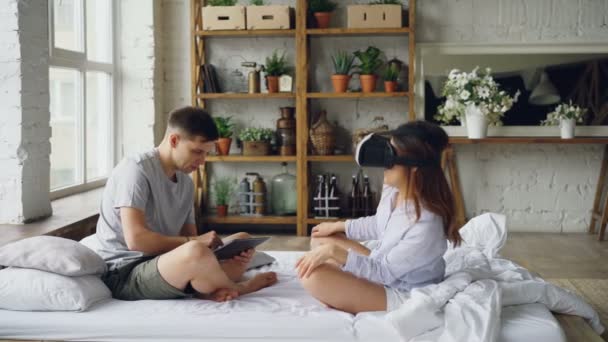 Married couple is having fun with augmented reality glasses, young woman is wearing then and smiling moving hands and her husband is using tablet. Technology and people concept. - Video, Çekim