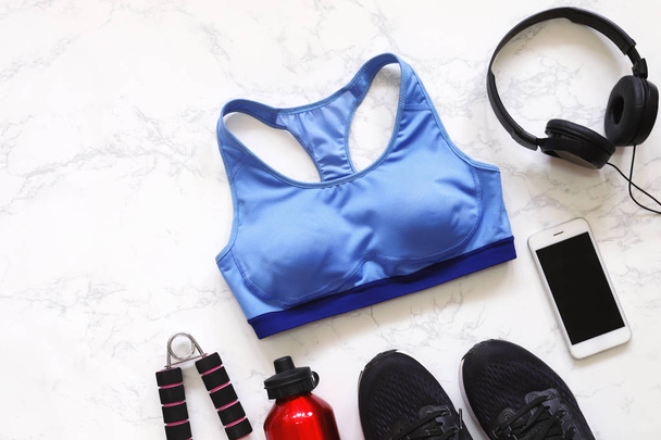 Top view of flat lay fitness equipment with sport bra, sneakers shoes, smartphone, earphones and bottle of water on white marble background - Photo, Image