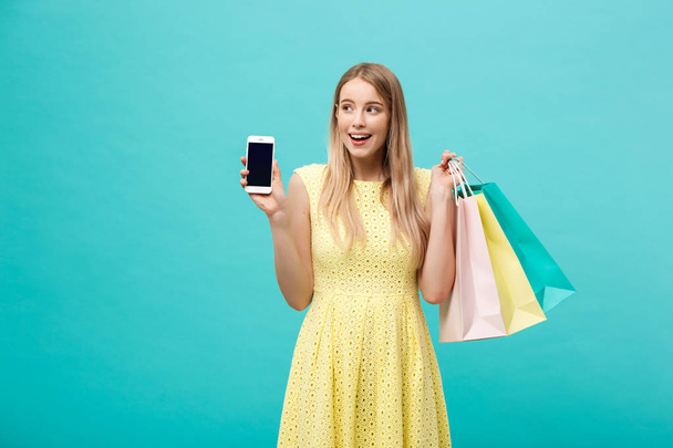 Portrait young attractive woman with shopping bags shows the phones screen directly to the camera. Isolated on blue background - Фото, зображення