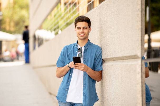 Outdoor shot of young male student in jeans denim clothes standing at street of modern European city and texting on smartphone - Foto, afbeelding