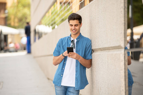 Outdoor shot of young male student in jeans denim clothes standing at street of modern European city and texting on smartphone - Photo, image