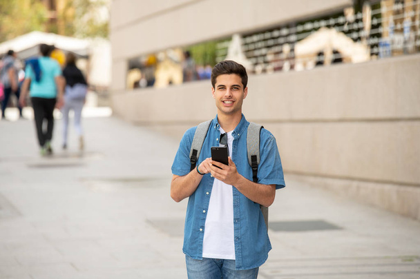 Outdoor shot of young male student in jeans denim clothes standing at street of modern European city and texting on smartphone - Fotografie, Obrázek