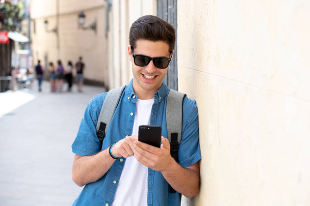 Outdoor shot of young male student in jeans denim clothes standing at street of modern European city and texting on smartphone - Foto, afbeelding