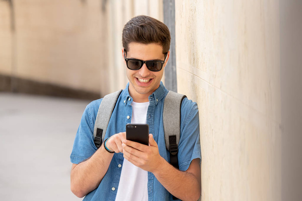 Tiro ao ar livre de jovem estudante do sexo masculino em jeans jeans roupas de pé na rua da cidade europeia moderna e mensagens de texto no smartphone
 - Foto, Imagem