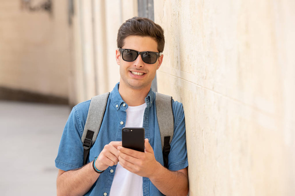 Outdoor shot of young male student in jeans denim clothes standing at street of modern European city and texting on smartphone - Fotografie, Obrázek