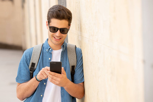 Tiro ao ar livre de jovem estudante do sexo masculino em jeans jeans roupas de pé na rua da cidade europeia moderna e mensagens de texto no smartphone
 - Foto, Imagem