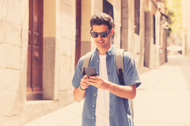 Outdoor shot of young male student in jeans denim clothes standing at street of modern European city and texting on smartphone - Foto, Imagen