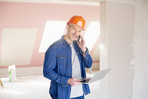 Uomo delle costruzioni sorridente fare chiamata e utilizzando il suo computer portatile mentre in piedi in cantiere
.  - Foto, immagini