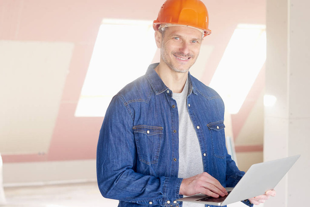 Работник среднего возраста держит ноутбук в руке, стоя на стройплощадке и работая
. - Фото, изображение