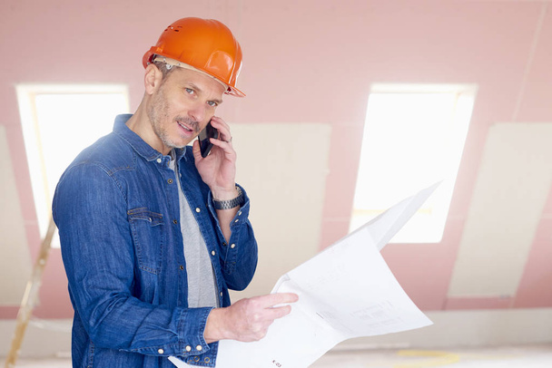 Middle aged construction worker holding plan in his hand while using mobile phone and talking with somebody. - Foto, Bild