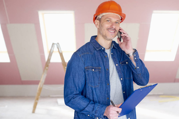 Middle aged handyman wearing hardhat and holding clipboard in his hand while while talking with somebody on mobile phone.  - Foto, Imagen