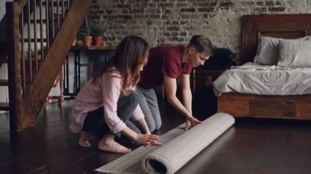 Young married couple is rolling out beautiful new carpet in bedroom at home then enjoying it. House interior, married life and happy people concept. - Materiał filmowy, wideo