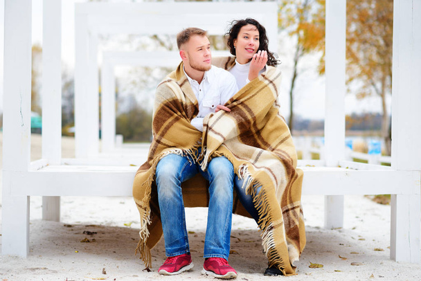 Casal jovem abraçando e coberto com um cobertor quente sentado na praia de outono
 - Foto, Imagem