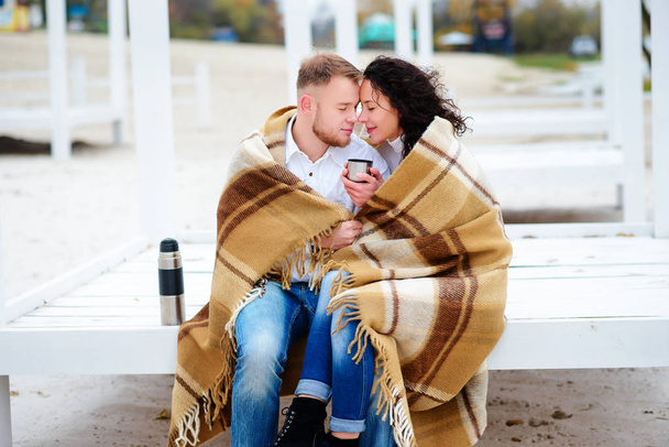 Young couple embracing and covered with a warm blanket sitting on autumn beach and drink coffee from a thermos - Photo, Image