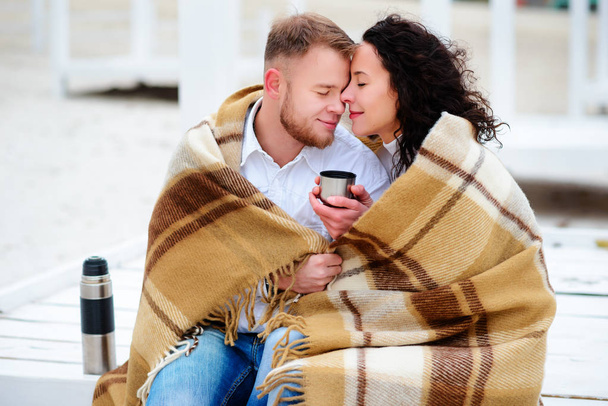 Νεαρό ζευγάρι αγκαλιάζει και να καλύπτεται με μια ζεστή κουβέρτα που κάθεται στο φθινόπωρο παραλία και πιείτε καφέ από ένα θερμός - Φωτογραφία, εικόνα