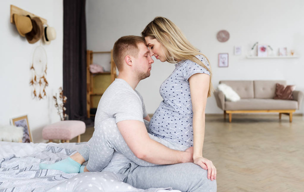 Side view of happy man and pregnant woman. They are sitting in bedroom with relaxation. - Foto, afbeelding
