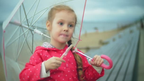 Little beautiful girl with umbrella playing in the rain eating ice cream on the coast - Séquence, vidéo