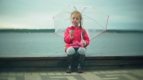 Krásná holčička s deštníkem v dešti jíst zmrzlinu na pobřeží - Záběry, video