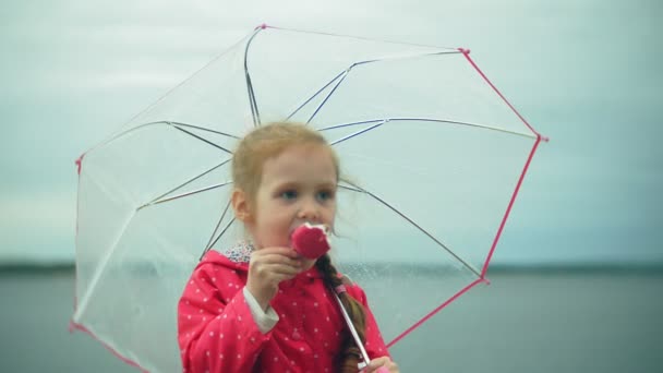 Pequena menina bonita com guarda-chuva jogando na chuva comendo sorvete na costa
 - Filmagem, Vídeo