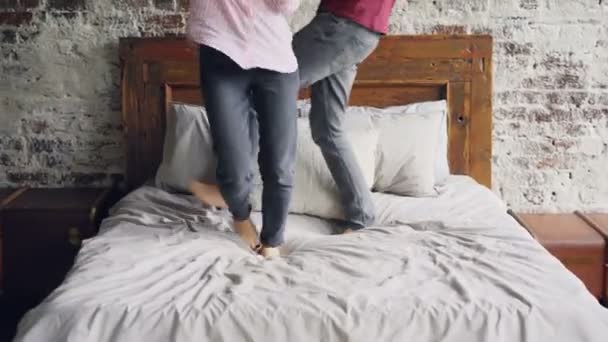 Happy lovers are dancing on bed holding hands and enjoying married life together. Modern double bed, brick wall and light loft apartment interior is visible. - Imágenes, Vídeo