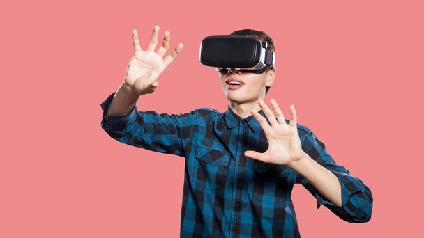 happy smiling young man wearing checkered shirt with vr headset on head trying to touch imaginary reality on pink background - Foto, afbeelding