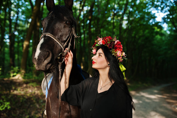 Mystical girl in wreath wear in black with horse in wood. - Photo, image
