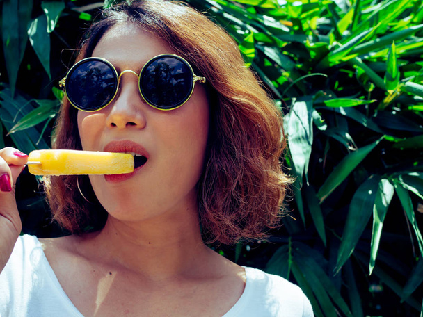 Όμορφη γυναίκα φορούσε άσπρο πουκάμισο και στρογγυλά γυαλιά ηλίου που τρώει κίτρινα Popsicle Pop πάγου τήξης στη θερινή ώρα με ηλιοφάνεια σε δέντρο φόντο κοντά μαλλιά - Φωτογραφία, εικόνα
