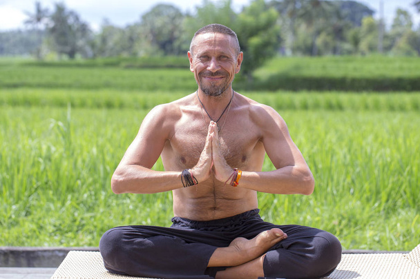 Man doing yoga and meditating in a position near a green rice terrace on the tropical island of Bali, Indonesia. Close up - Fotografie, Obrázek