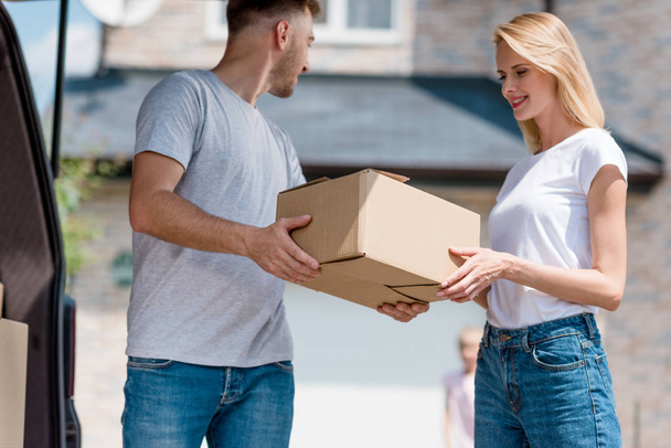 Мужчина отдает картонную коробку жене, пока их дочь стоит во дворе нового дома
  - Фото, изображение