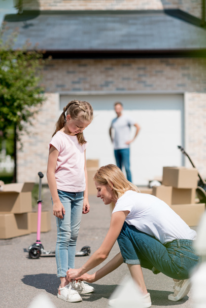 side view of smiling woman tying shoelaces of daughter and man standing behind near cardboard boxes in yard of new cottage  - Photo, image