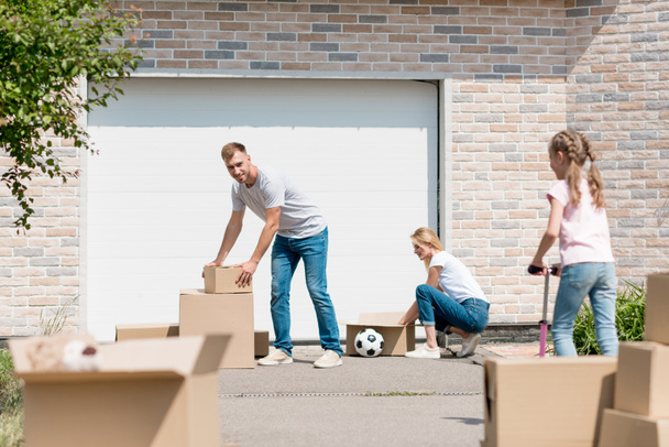 young couple unpacking cardboard boxes while their daughter riding on kick scooter in front of new cottage  - Photo, Image