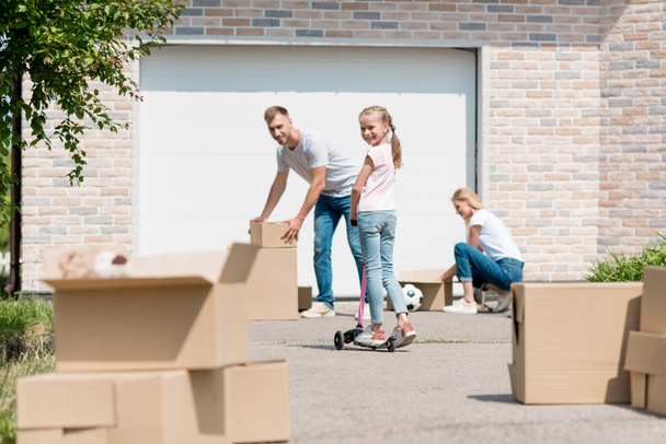 couple unpacking cardboard boxes while their daughter riding on kick scooter in front of new cottage  - Photo, Image
