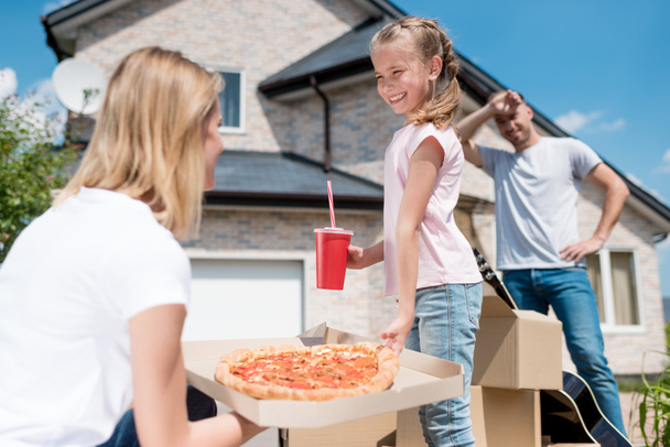 side view of happy mother holding box with pizza and daughter with cup of cola taking slice of pizza while father standing behind of new house  - Photo, Image