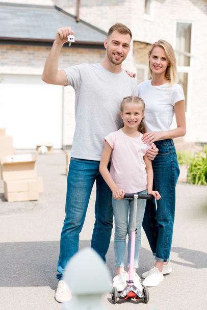 man holding key with trinket while his wife embracing daughter on kick scooter in front of new house  - Foto, Imagem
