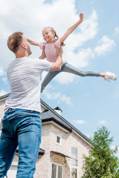 Tiefansicht des Vaters, der lächelnde Tochter mit breiten Armen vor dem Haus aufzieht  - Foto, Bild