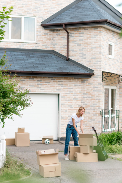 young woman unpacking cardboard boxes near guitar in front of new cottage  - Photo, Image