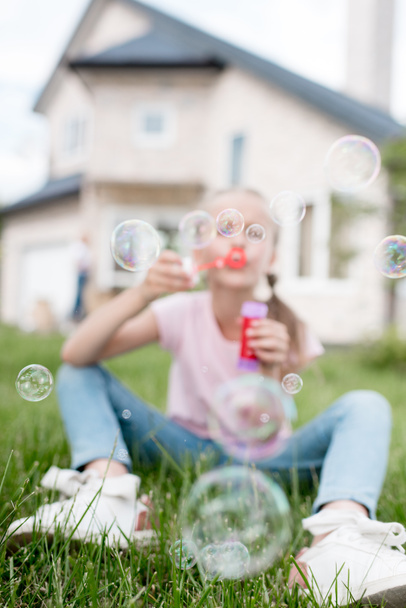 Kleines Kind mit Seifenblasen sitzt auf dem Rasen, während die Mutter hinten vor dem Haus steht  - Foto, Bild