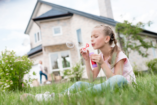 Seitenansicht eines kleinen Kindes, das Seifenblasen pustet und auf dem Rasen sitzt, während seine Mutter hinter dem Haus steht  - Foto, Bild
