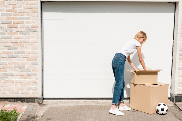 smiling young woman unpacking cardboard boxes near soccer ball in front of garage of new house  - Foto, Imagen