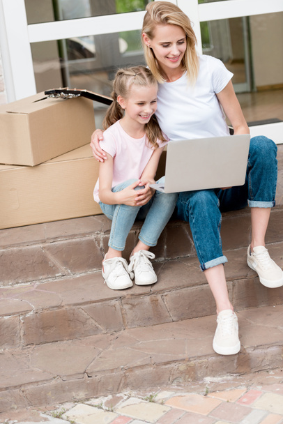 happy woman with daughter using laptop on stairs with cardboard boxes and guitar in front of new house  - Photo, Image