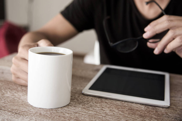close-up de um jovem caucasiano, sentado a uma mesa, usando um tablet com uma xícara de café em uma mão e um par de óculos na outra mão
 - Foto, Imagem