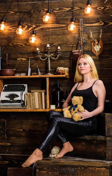 Girl in black clothes holds teddy bear toy in hand, wooden interior on background. Woman on dreamy face relaxing in wooden interior. Lady blonde enjoy leisure with teddy bear. Rest and relax concept - Фото, изображение