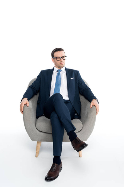 full length view of serious middle aged businessman in eyeglasses and formal wear sitting in armchair and looking away on white - Zdjęcie, obraz
