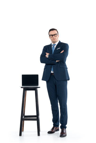 confident businessman standing with crossed arms near laptop on stool on white - Фото, изображение