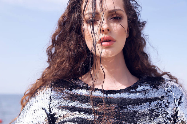 fashion outdoor photo of beautiful girl with dark curly hair in elegant shiny dress with sequins posing on summer beach - Φωτογραφία, εικόνα